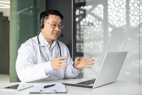 Physician in a lab coat talking on a laptop.